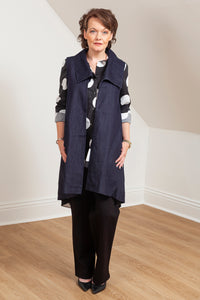 Woman wearing a navy blue vest (Melva - Front view) over a patterned shirt in a room with white walls and wooden floor. 