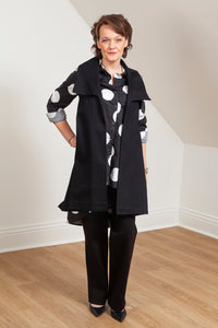 Woman wearing a black vest (Melva - Front view) over a patterned shirt in a room with white walls and wooden floor. 