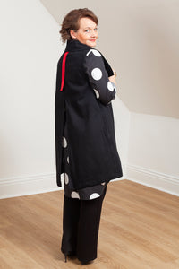 Woman wearing a black vest (Melva - side /back view) over a patterned shirt in a room with white walls and wooden floor. 
