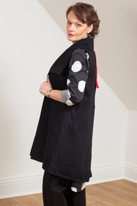Woman wearing a black vest (Melva - side view) over a patterned shirt in a room with white walls and wooden floor. 
